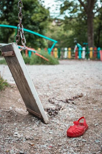 Kırık salıncak ve kırmızı çocuk sandaleti kumda zincirle yatıyor. Üzüntü, kayıp çocuklar günü konsepti