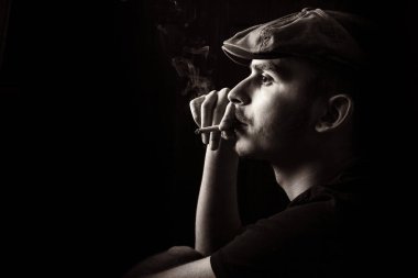 Siyah beyaz, siyah arka planda sigara içen, bıyıklı, siyah şapkalı hoş bir gencin portresi..