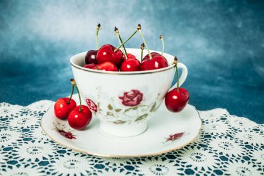 Masanın üzerinde, altında tığ işi olan porselen bir fincanda kırmızı, lezzetli kirazlar. Mavi arkaplan