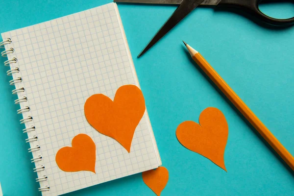 Cuaderno, lápiz, tijeras y corazones de color naranja sobre un fondo azul . — Foto de Stock
