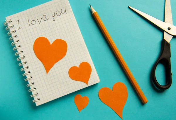 Cuaderno, lápiz, tijeras y corazones de color naranja sobre un fondo azul . — Foto de Stock
