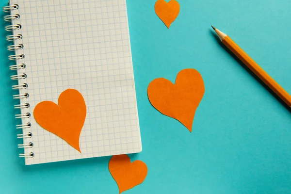 Bloc de notas, lápiz y corazones de color naranja sobre un fondo azul . — Foto de Stock