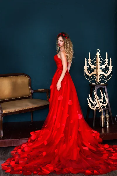 Beautiful young blonde woman in red long dress with petals on a black background. queen, red hoop on the head, golden curls, passionate girl in red, woman with red lips, princess from a fairy tale, — Stock Photo, Image