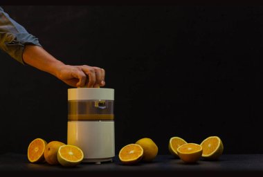 Portakal sıkan bir el, siyah arkaplanlı portakal suyu ve daha fazla bölünmüş portakal hazırlamak için makinede çekilmiş.