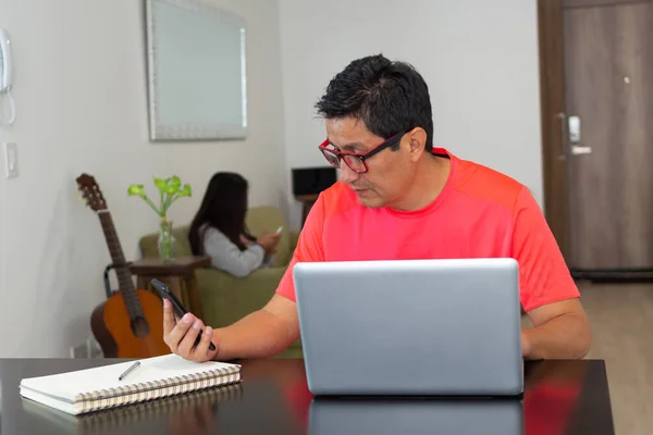 In the living room of the house a man works with his cell phone and laptop while in the living room his daughter is looking at messages