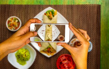 Cep telefonuyla odaklanmış eller Taco, chili ve guacamole ile bir tabak çözülmüş Meksika yemeğine fotoğraf çekiyor.