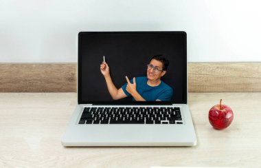 Öğretmen, bir karatahtanın önünde bilgisayar monitörünün ekranında bir masada yanında elma olan bir bilgisayar ekranında ders veriyor.