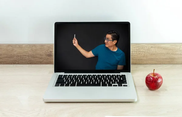 Virtual class taught by a teacher on a laptop screen smiling as he writes on a black board with an apple on the desk