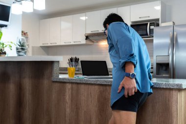 İç çamaşırlı ve gömlekli bir adam video konferansı düzenliyor. Evinin mutfağında bilgisayarını bembeyaz gösteriyor.