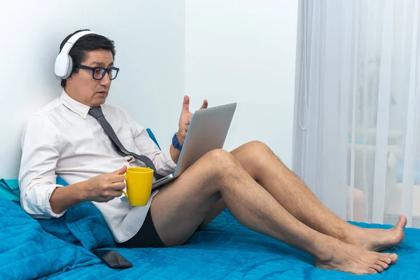 Man with headphones and a cup of coffee talks on the internet with his computer from the bedroom of his house