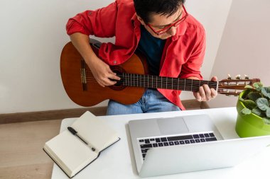Evdeki bir odada bir adam online ders sırasında dizüstü bilgisayarıyla gitar çalar.