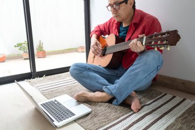 İnternet dersinde klasik gitarını çalan bir adam evdeki bir halıda oturuyor.