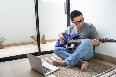 Evdeki bir odada halının üzerinde oturan bir adam elektro gitar dersleri alıyor.