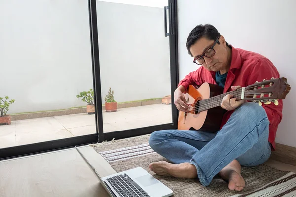 Online acoustic guitar classes. A man plays his guitar barefoot and relaxed on the carpet in a room at home