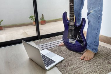 Elektro gitar çevrimiçi dersi. Evdeki bilgisayarın önündeki paspasın üzerinde çıplak ayakla duran bacaklar.