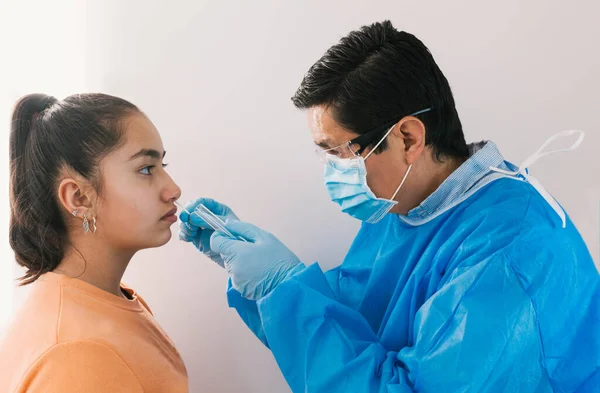 Maskeli, gözlüklü, koruyucu elbiseli ve eldivenli bir doktor genç bir kızdan koronavirüs ya da solunum hastalığı muayenesi için burun örneği alıyor.