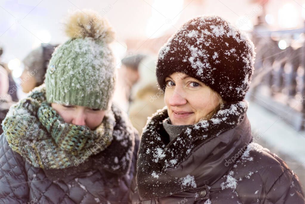 Two girls in warm winter clothes in heavy snow. Brightly visited city street. Snowflakes lie on their clothes and hats.