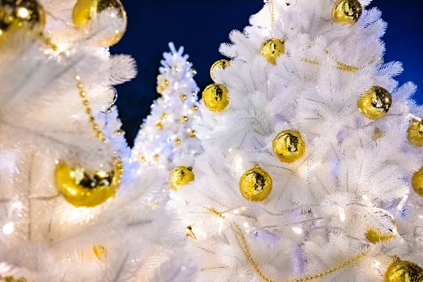 Ярко освещенные белые искусственные елки ночью. Золотые шары, покрытые каплями воды, и гирлянды, свисающие на белых ветвях. Новогодние атрибуты большого города создают праздничное настроение . — стоковое фото