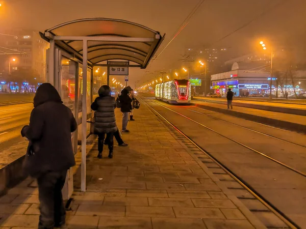 Moscow.Russia. December 26, 2019. Trams in the fog lit by a network of LEDs arrive at a bus stop with passengers. Environmentally friendly public transport, decorated in honor of the New Year holidays — Stock Photo, Image