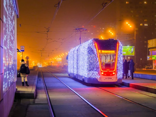 Moscow.Russia. December 26, 2019. Trams in the fog lit by a network of LEDs arrive at a bus stop with passengers. Environmentally friendly public transport, decorated in honor of the New Year holidays — Stock Photo, Image