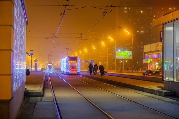 Русскость 26 декабря 2019 года. Трамваи в тумане, освещенные сетью светодиодов, прибывают на автобусную остановку с пассажирами. Экологически чистый общественный транспорт, украшенный в честь новогодних праздников — стоковое фото