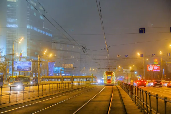 モスクワ、ロシア。2019年12月26日道路網によって照らされたトラムが霧の多い夜にレールを走行します。環境に優しい公共交通機関、新年の休日を記念して飾られた. — ストック写真
