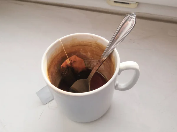 在未完成的茶的残渣中 一个湿的用过的一次性茶袋躺在脏兮兮的瓷杯底部 杯子里有一个金属茶匙 红茶花斑长杯的内壁 — 图库照片