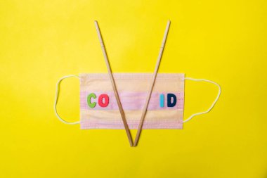 Sarı arka planda pembe tıbbi maske ve V harfi şeklinde Asya yemek çubukları. Yazıya göre virüs. Asya 'dan bir tedavi olarak hastalıklar.