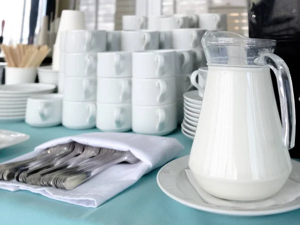 A glass jug with milk on the table. Nearby are many stainless steel forks. In the background are stacks of white ceramic cups and saucers. Preparing for tea and coffee in a restaurant.