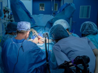 A group of surgeons performing minimally invasive surgery on the patient's anus using surgical instruments. Surgeons have sterile blue surgical clothing.