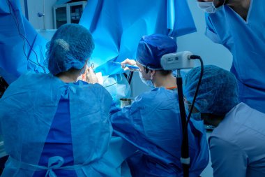 A group of surgeons performing minimally invasive surgery on the patient's anus using surgical instruments. Surgeons have sterile blue surgical clothing.