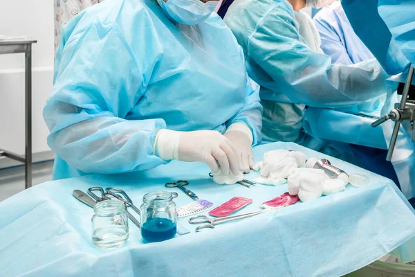 Sterile surgical instruments and glass containers with solutions on the table during a surgical operation. Above the table are the hands of a surgeon in latex gloves.