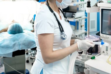 Beyaz tıbbi giysiler içinde bir anestezi uzmanı, boynunda bir maske ve steteskop ile bir hastayı ameliyata hazırlıyor. Yüksek teknolojili cerrahi ekipmanların yanında. Hasta ameliyat masasında yatıyor..