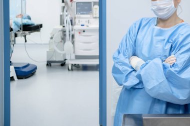 Ameliyat kıyafetleri, maske ve steril eldiven giymiş bir kadın cerrah kollarını ameliyathanenin girişinde kavuşturmuş bekliyor. Ameliyathanede, masanın üstünde hasta var.. 