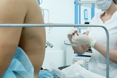 Beyaz tıbbi giysiler içinde bir anestezi uzmanı, bir maske ve steril eldiven elinde bir iğne tutuyor. Epidural anestezi uygulanacak olan hastanın arkası. Ameliyat için hazırlanıyor..