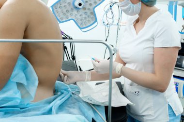 Beyaz tıbbi giysiler içinde bir anestezi uzmanı, bir maske ve steril eldiven elinde bir iğne tutuyor. Epidural anestezi uygulanacak olan hastanın arkası. Ameliyat için hazırlanıyor..