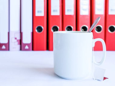 Ofiste çay. Belgeler için kırmızı ofis klasörlerinin arkasında çay olan beyaz seramik bardak. Tek kullanımlık poşetlerden çay. Bir çay kaşığı fincanda duruyor. İşte çay molası..