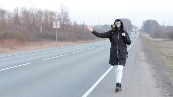 Dívka v plynové masce stojí na kraji silnice a snaží se zastavit auto. — Stock video