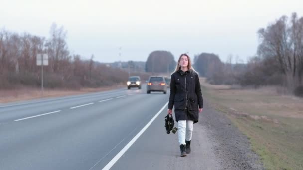 En svart gasmask sveper i handen på en flicka som går längs kanten av en motorväg — Stockvideo