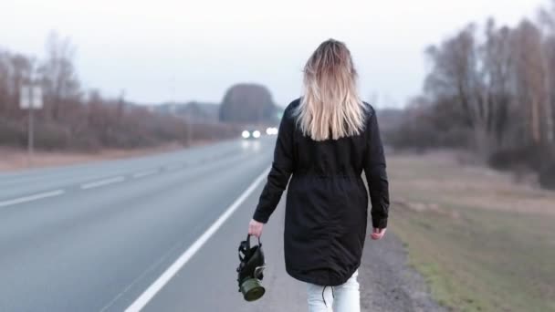 En svart gasmask sveper i handen på en flicka som går längs kanten av en motorväg — Stockvideo
