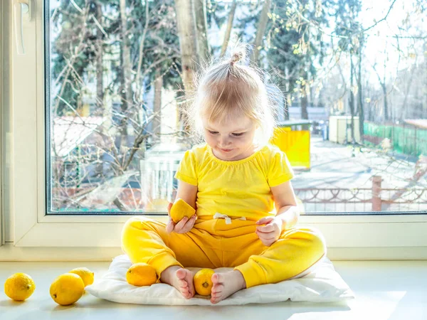 A little girl in yellow clothes sits on a window sill during the day and smiles. Girl holding lemon legs. A bright sun shines in the back of the girl. Lemons as a disease prevention.
