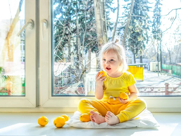 A little girl in yellow clothes with a lemon in her hands sits on a windowsill in the afternoon and smiles. The bright sun shines. Useful citrus fruits as prevention of viral and respiratory diseases.