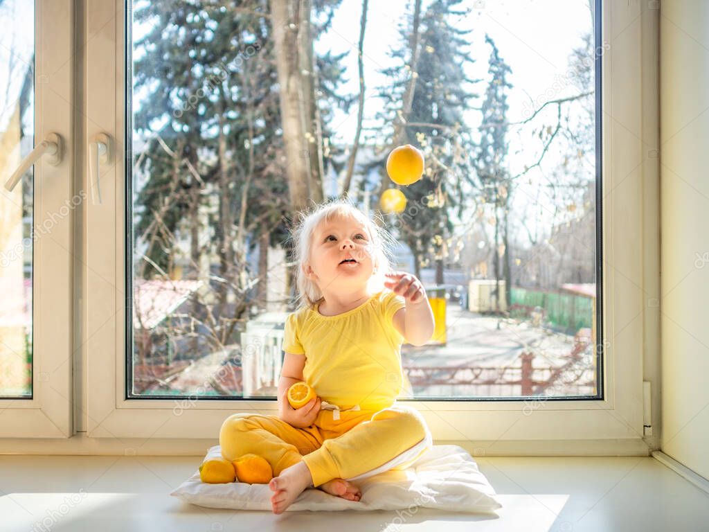 Little girl in yellow clothes sits on a windowsill in the afternoon. She throws up a lemon and laughs. The bright sun shines in her back. Lemons as a prevention of diseases and strengthening immunity