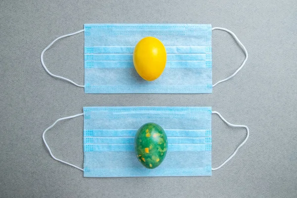 Yellow and green easter eggs lie on blue medical masks on a gray background. Flat lay. Easter holiday concept during quarantine coronavirus epidemic. Copy space