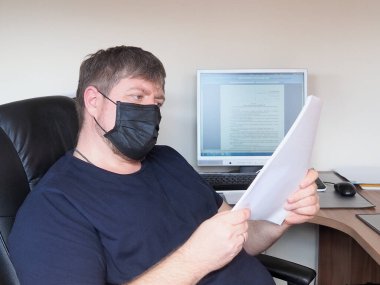Siyah tıbbi maskeli yetişkin bir adam, bilgisayarın önündeki sandalyede oturan belgeleri dikkatle inceler. Coronavirüs salgınının karantinası sırasında ofis işi kavramı.