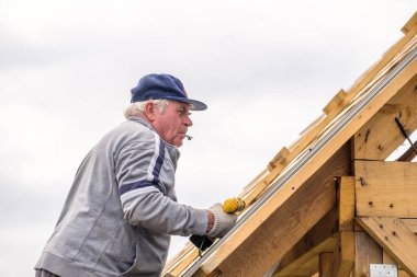 Kıdemli inşaat işçisi tornavida kullanıyor, inşaat halindeki bir kır evinin çatısındaki ahşap kirişlere çatı örtüsü bağlıyor. Yaşlıların fiziksel aktiviteleri.