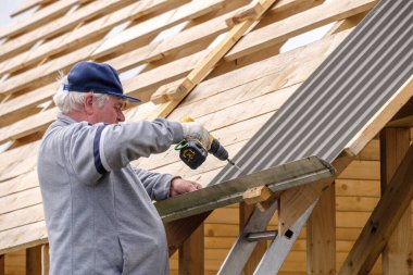 Kıdemli inşaat işçisi tornavida kullanıyor, inşaat halindeki bir kır evinin çatısındaki ahşap kirişlere çatı örtüsü bağlıyor. Yaşlıların fiziksel aktiviteleri.