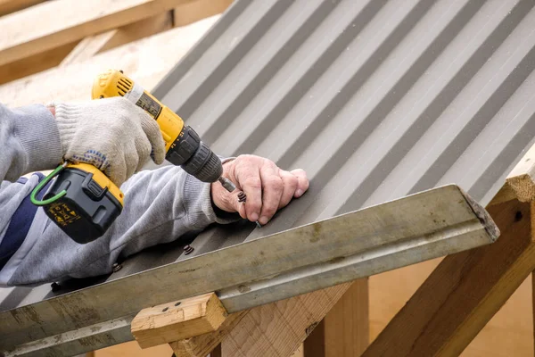 Male hands in work gloves with a yellow screwdriver screw the roofing sheet to the roof of the country house. Cordless drill.