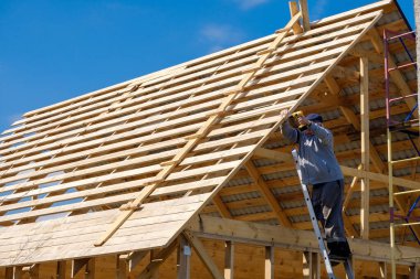 Kıdemli gri saçlı müteahhit mavi gökyüzüne karşı bir bahar günü kablosuz tornavidayla ahşap bir kır evinin çerçevesini topluyor. Yaşlıların fiziksel aktiviteleri. Çevre dostu konutlar.