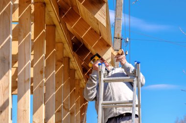 Kıdemli gri saçlı müteahhit mavi gökyüzüne karşı bir bahar günü kablosuz tornavidayla ahşap bir kır evinin çerçevesini topluyor. Yaşlıların fiziksel aktiviteleri. Çevre dostu konutlar.